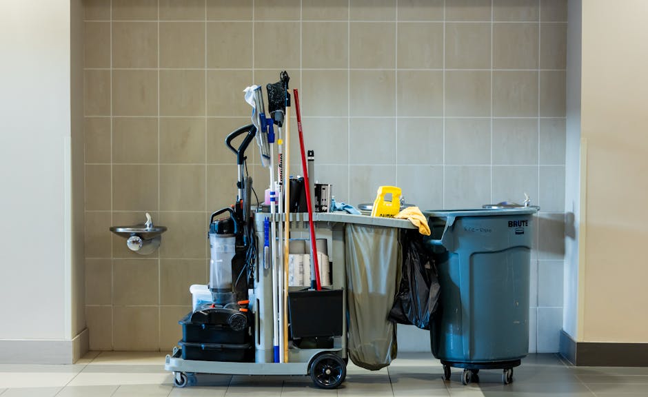 A cleaning trolley situated against a beige tiled wall in a commercial office setting, with various cleaning tools including mops, brushes, and spray bottles arranged on the trolley. To the right, the