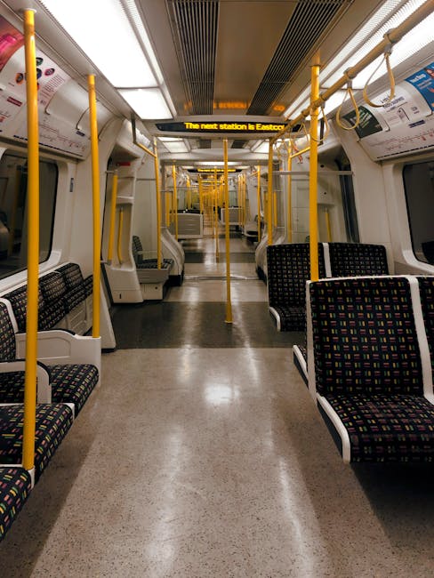 Interior view of a train carriage with clean, well-maintained fabric-upholstered seats in a black and colorful pattern, arranged in pairs along the sides of the aisle. The train features yellow handra